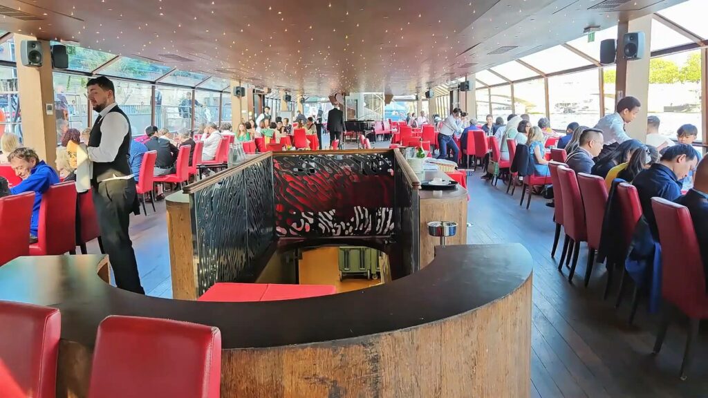 Salle à manger élégante avec tables dressées, baies vitrées montrant Paris illuminé, et musiciens en fond